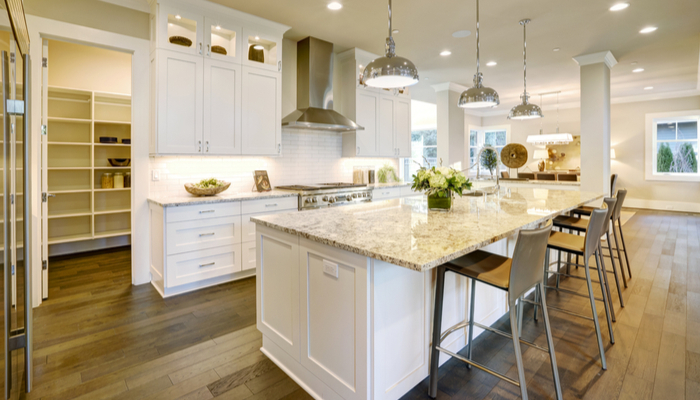 Bright white kitchen design with wooden floor features large bar style kitchen island with granite countertop and has an open door lead to walk-in pantry