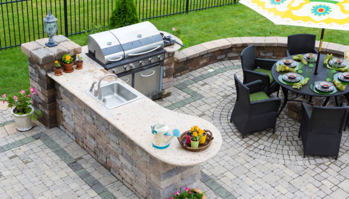 High angle view of a stylish outdoor kitchen, gas barbecue and dining table set for entertaining guests with formal place settings and flowers on a paved patio with chairs and patterned umbrella.jpg