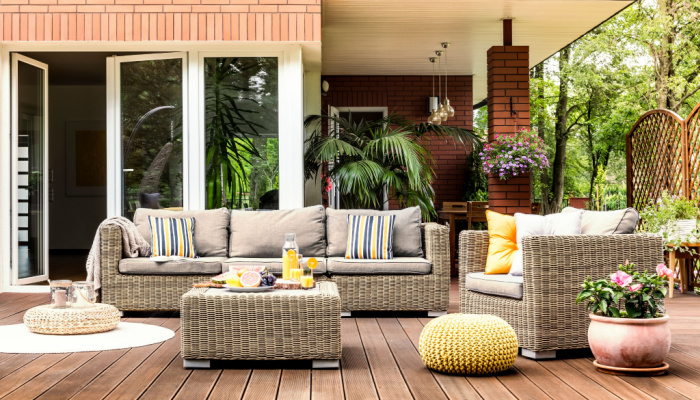 Yellow pouf next to a rattan armchair and flowers on wooden patio with striped pillows on sofa and plants and flowers