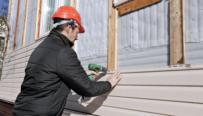 A worker on a black jacket with red helmet installs panels beige siding on the facade of the house in the morning