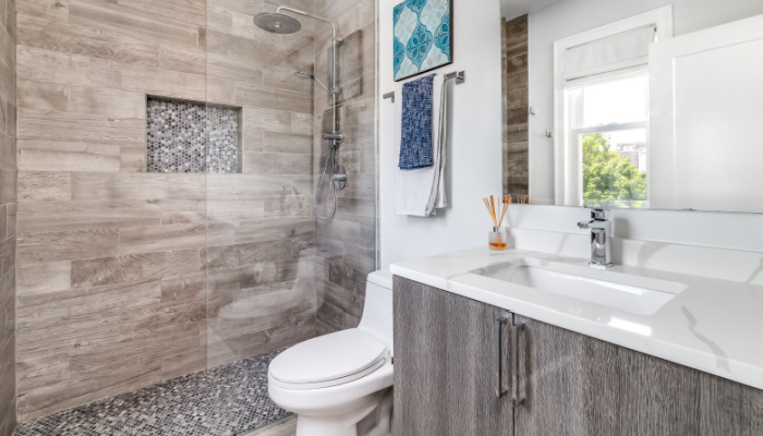 A small bathroom with a white marble counter top, wood cabinet, and a custom tiled shower with a shelf picture taken in daylight