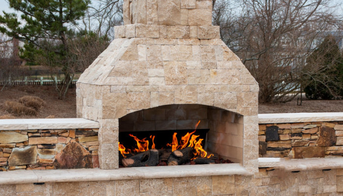 A gas fireplace made from paving stone burning outside on a patio close up with trees in the backgroud during fall season