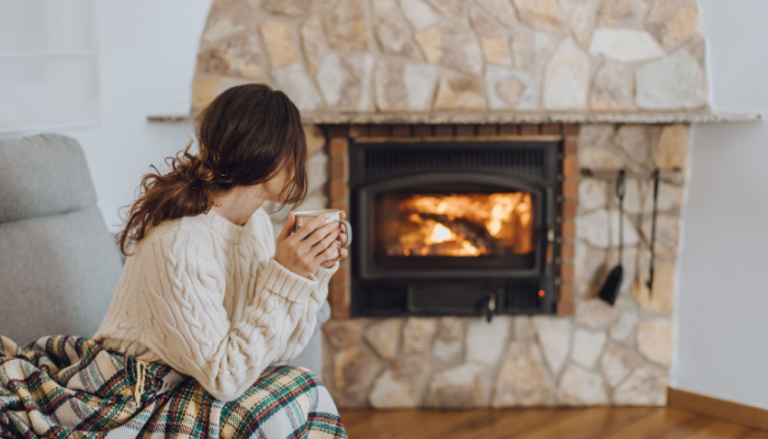 Young woman wearing white woollen sweater sitting at home by the fireplace with a hot tea or coffee mug and warming her hands